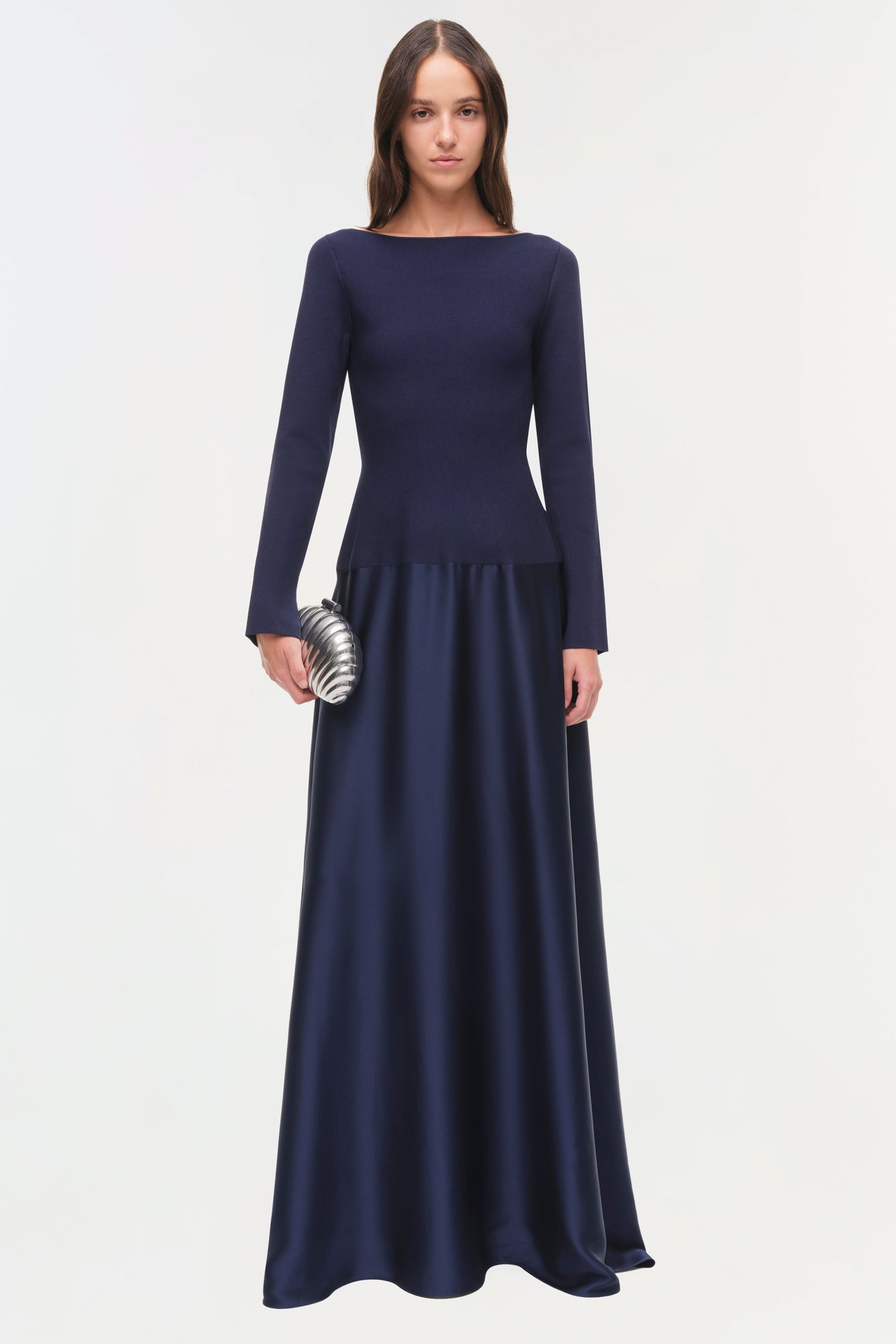 Woman wearing navy satin knit gown with long sleeves, fitted bodice, and flowing skirt to floor, holding metallic clutch.