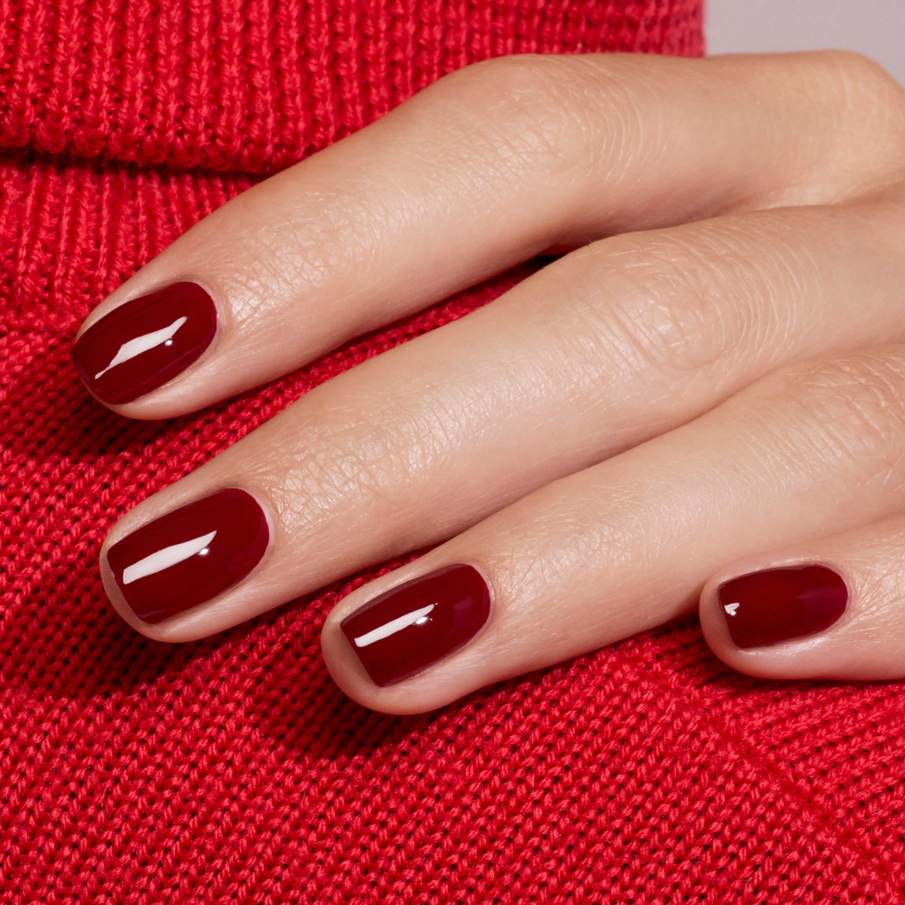 A hand with neatly manicured nails painted in Manucurist’s Dark Pansy, a glossy burgundy shade, rests on red knit fabric, perfectly matching the velvet red sweater underneath.