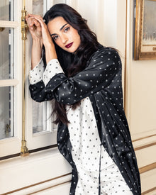 A woman with long dark hair and bold lipstick stands by a window in an elegantly decorated room, wearing Petite Plumes Womens Silk Cami Short Set in White Art Nouveau, reminiscent of an Art Nouveau print. She gazes thoughtfully, leaning against the wall.