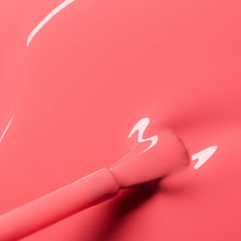 A close-up of a brush applying Manucurist's Capucine gel polish, a glossy coral pink shade with reflections of light shimmering on its smooth, wet surface.