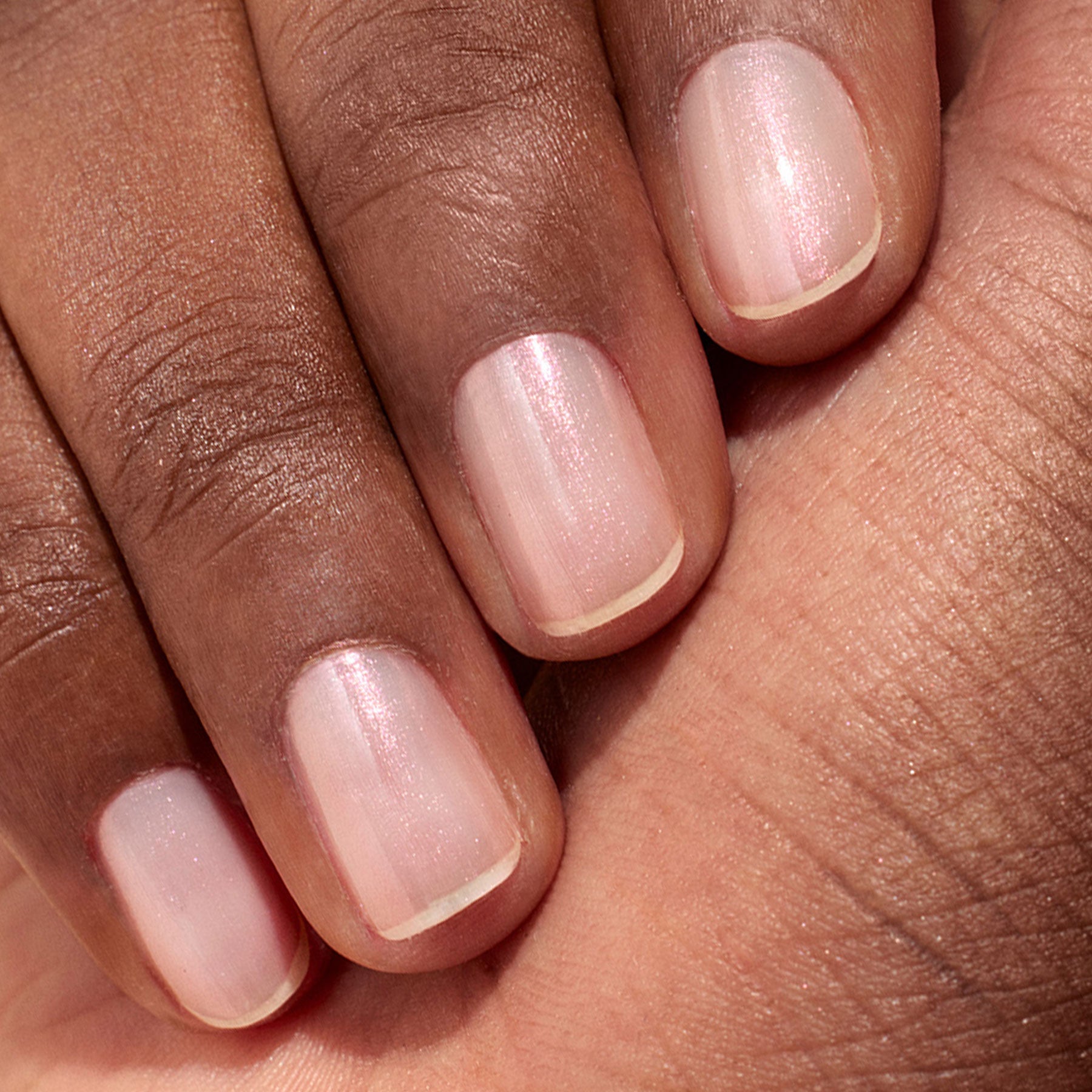 A close-up of a hand with medium brown skin, showcasing neatly manicured nails painted in Manucurist's Glitters Duo Rose Gold  Glazed—a sheer pale pink polish with a delicate pearlescent shine.