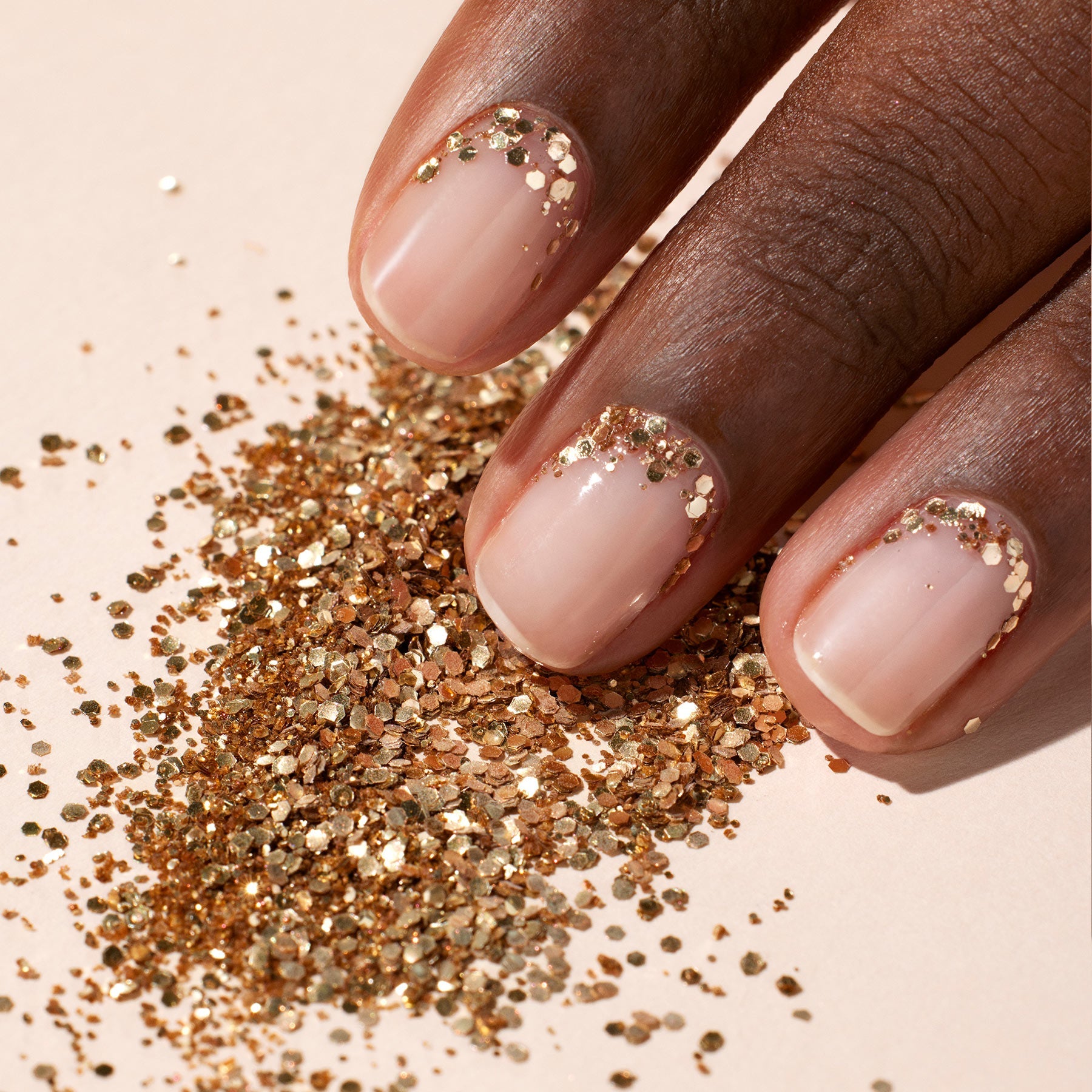 A hand with nude nails sparkles at the cuticles with Manucurist's Glitters Duo Rose Gold  Glazed, mirroring loose gold glitter scattered on a light surface beneath the fingers.