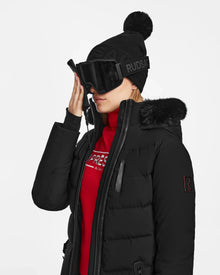 Wearing the Rudsak ALAIA - 8124816 Black womens quilted ski jacket with a fur-trimmed hood, black ski goggles, and a black pom-pom beanie, a person adjusts their glasses against a plain white background.