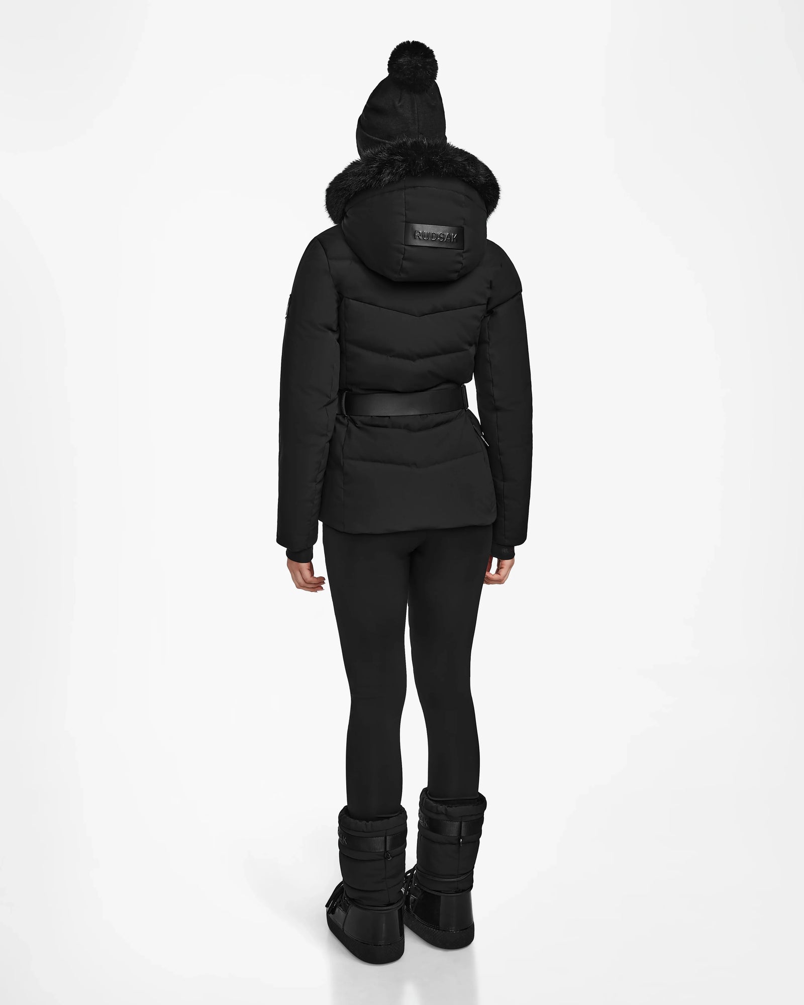 A person faces away against a white background, wearing the Rudsak ALAIA - 8124816 Black womens quilted ski jacket with fur-trimmed hood, paired with black leggings, boots, and a black knit hat with a pom-pom.