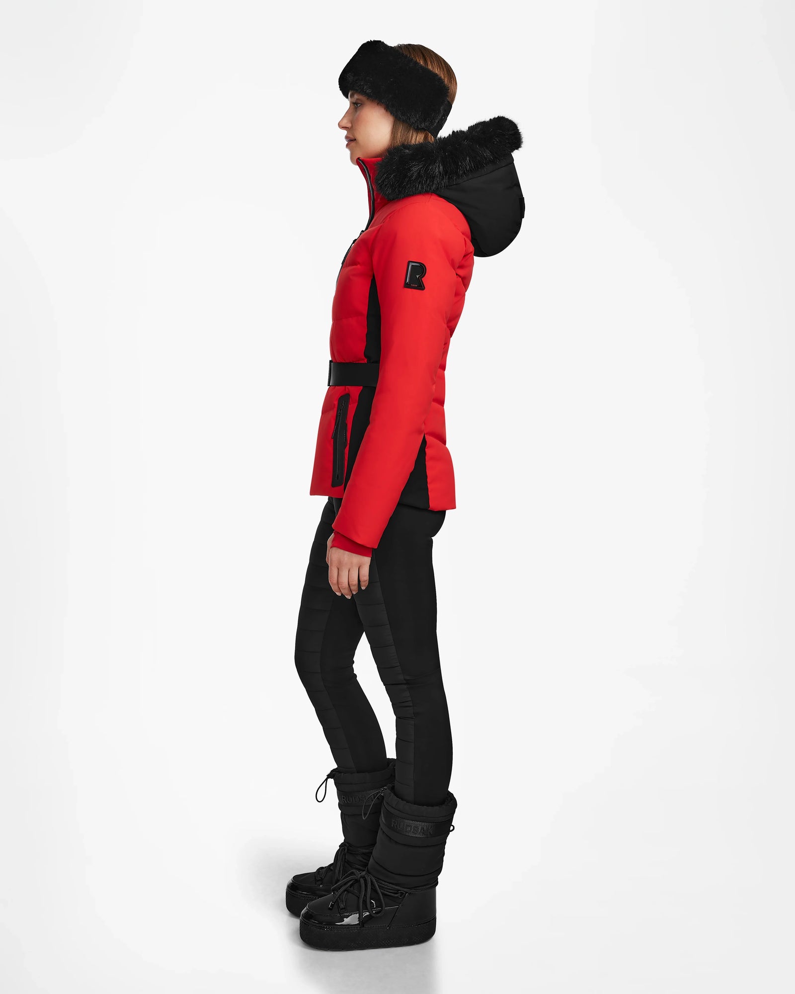 A person stands in profile against a white background wearing the Rudsak ALAIA - 8124816 Flame Red ski jacket, styled with black leggings, winter boots, and a black headband.