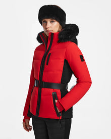 A woman in the Rudsak ALAIA - 8124816 Flame Red quilted ski jacket with a fur-trimmed hood and black fur hat stands against a plain white background, gazing slightly to the side.