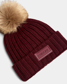 The ANISA - L423500 Wine by Rudsak is a ribbed, burgundy knit beanie with a large faux fur pom pom and folded brim featuring a rectangular Rudsak patch—an essential women’s winter accessory.