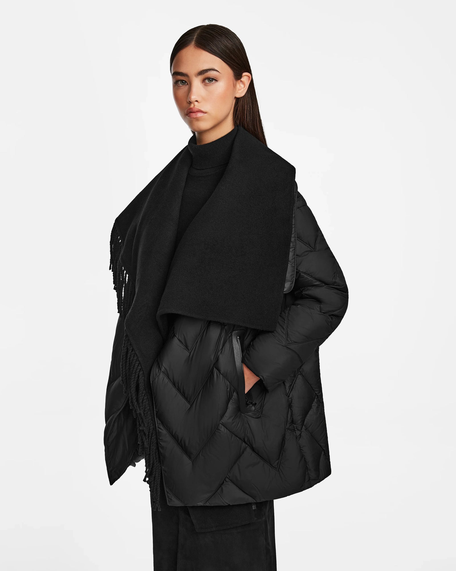 A woman with long dark hair wears the Rudsak ANNE MARIE - 8124713 Black mid-length puffer jacket, styled with a large scarf and pants, standing against a plain white background and looking at the camera with a neutral expression.