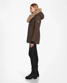 A woman in profile wears the Rudsak AURORE - L123561 Espresso fitted down parka with a fur-trimmed collar, paired with black leggings and ankle boots, standing against a plain white background.