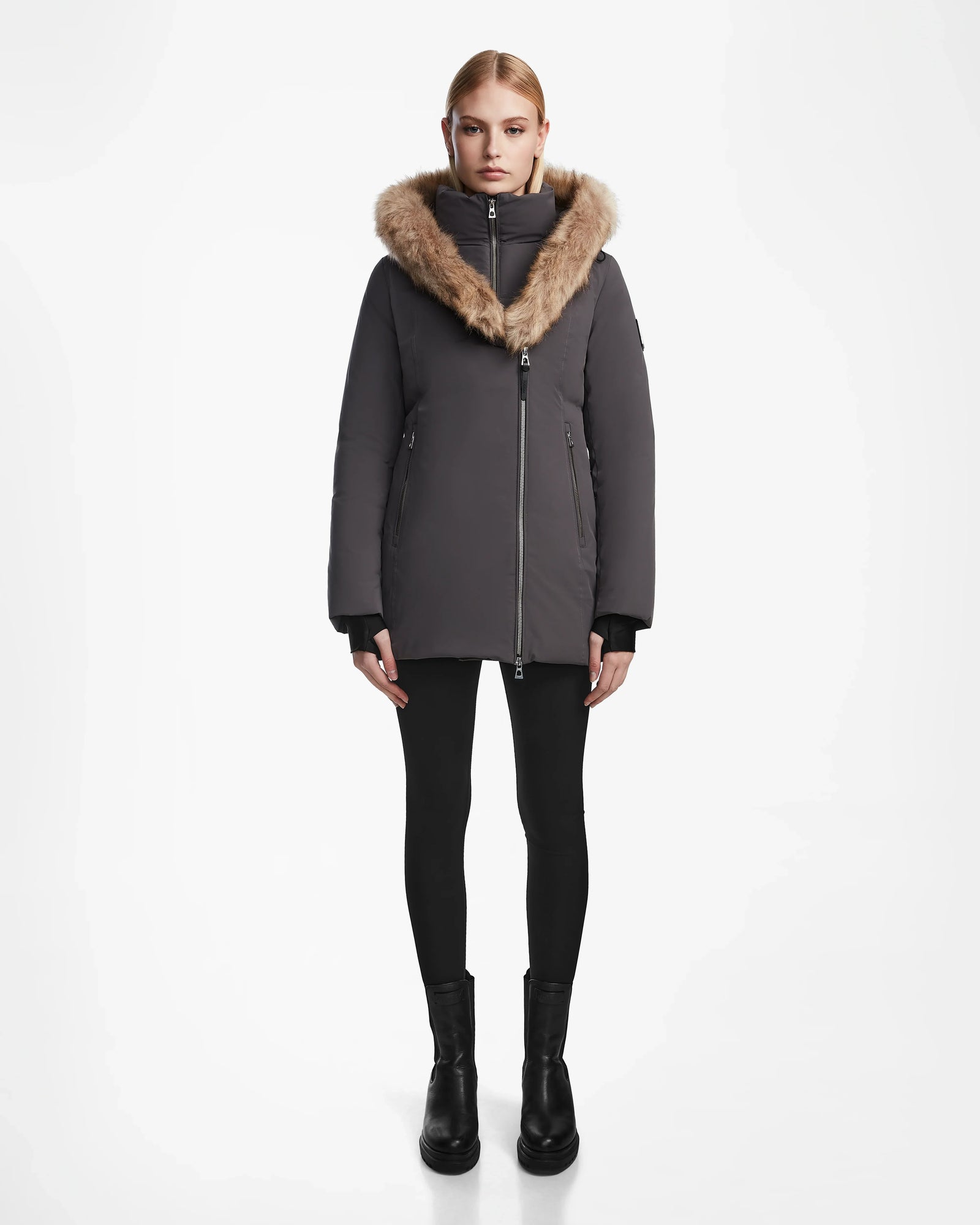 A woman faces forward against a plain white background, wearing the Rudsak AURORE - L123561 Grey fitted winter coat with a large fur-trimmed hood, paired with black leggings and black ankle boots.