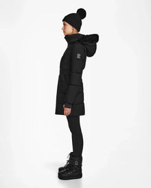 A person in profile wears the Rudsak CHRISTEL M-M - L124505 Black quilted winter coat with a fur-trimmed hood, styled with black leggings, winter boots, and a black knit beanie with pom-pom against a white background.
