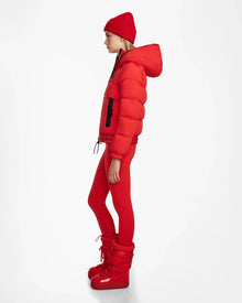 A person stands in profile against a white background, wearing the Rudsak DANI M - 8124539 Flame Red après-ski jacket styled with red leggings, beanie, and boots for a sleek and sporty monochromatic look.