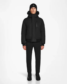 A person faces forward against a white background, wearing the Rudsak DEACON - M123513 Black/Black mens winter coat with a fur-lined hood, paired with black pants, shoes, and a beanie.
