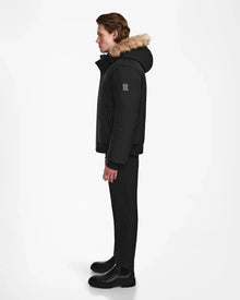 A person stands in profile facing left, wearing the Rudsak DEACON - M123513 Black/Natural down puffer jacket with fur trim, paired with black pants and boots against a plain white background.