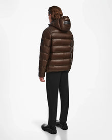 A man faces away wearing the Rudsak DEMIAN S - M123508 Espresso, a mens shiny dark brown water- and wind-resistant down puffer, paired with black pants and shoes, set against a plain white background.