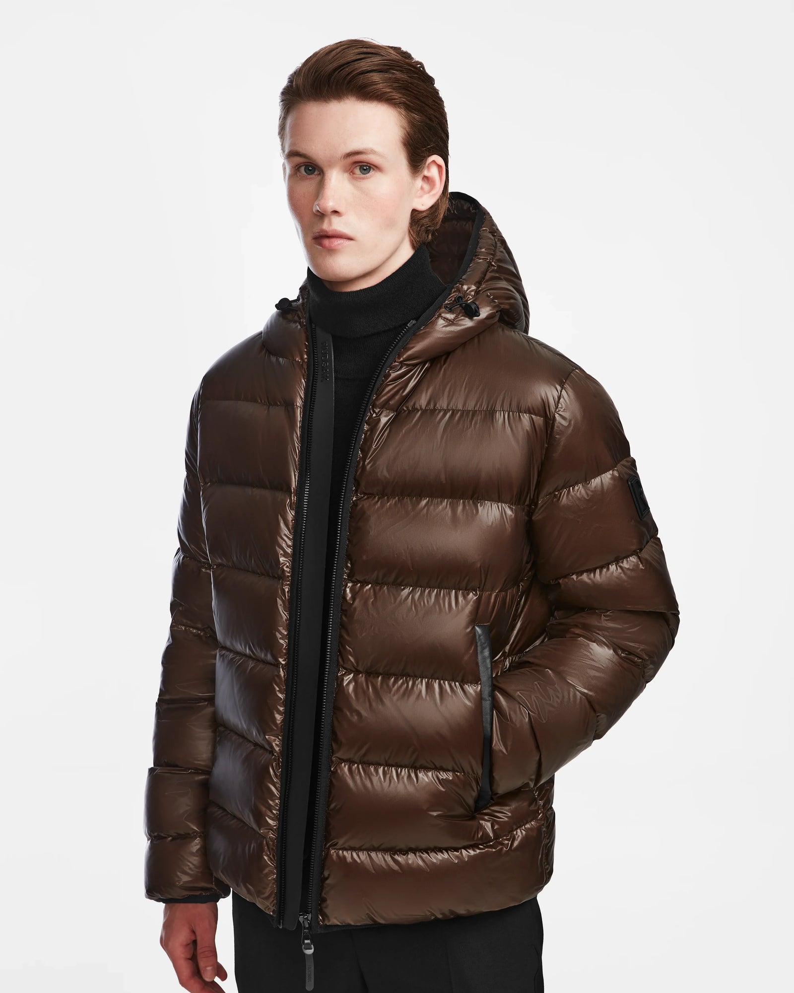 A young person models the Rudsak DEMIAN S - M123508 Espresso men’s down puffer jacket, with premium down fill, black turtleneck, and pants, standing against a plain white background and looking directly at the camera.