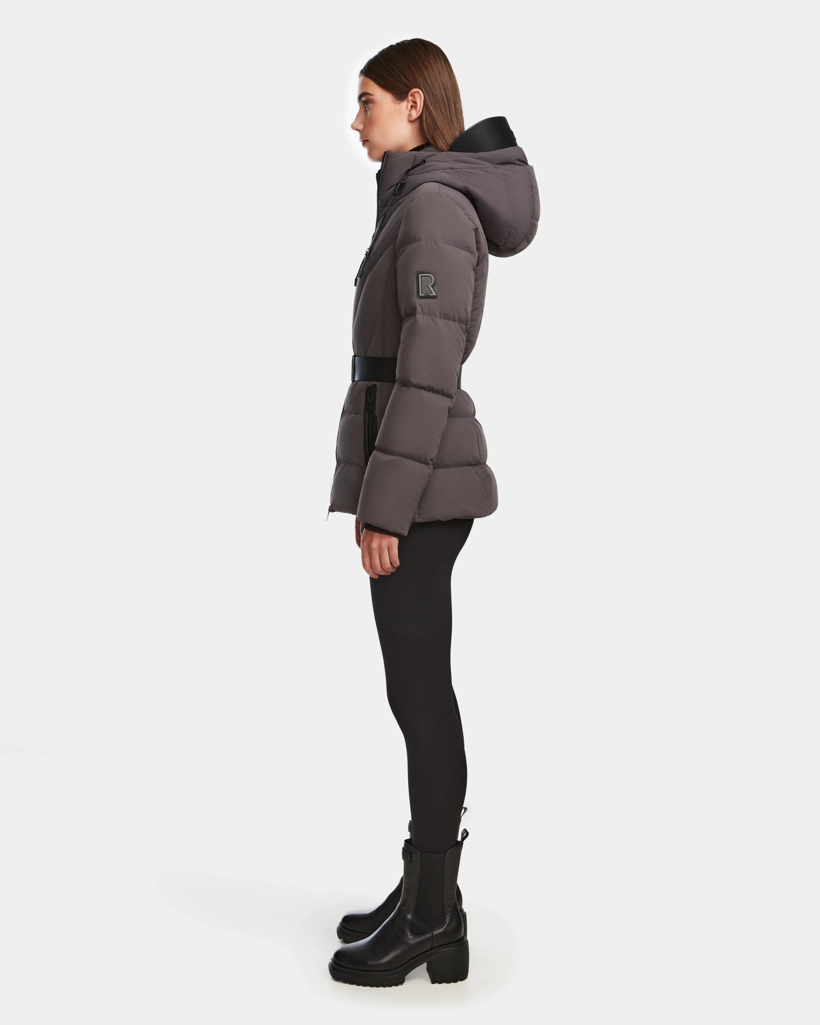 A woman stands in profile wearing the Rudsak ENZAH - 8124692 Grey RDS-certified down women’s puffer jacket with a black belt, black leggings, and ankle boots against a plain white background.