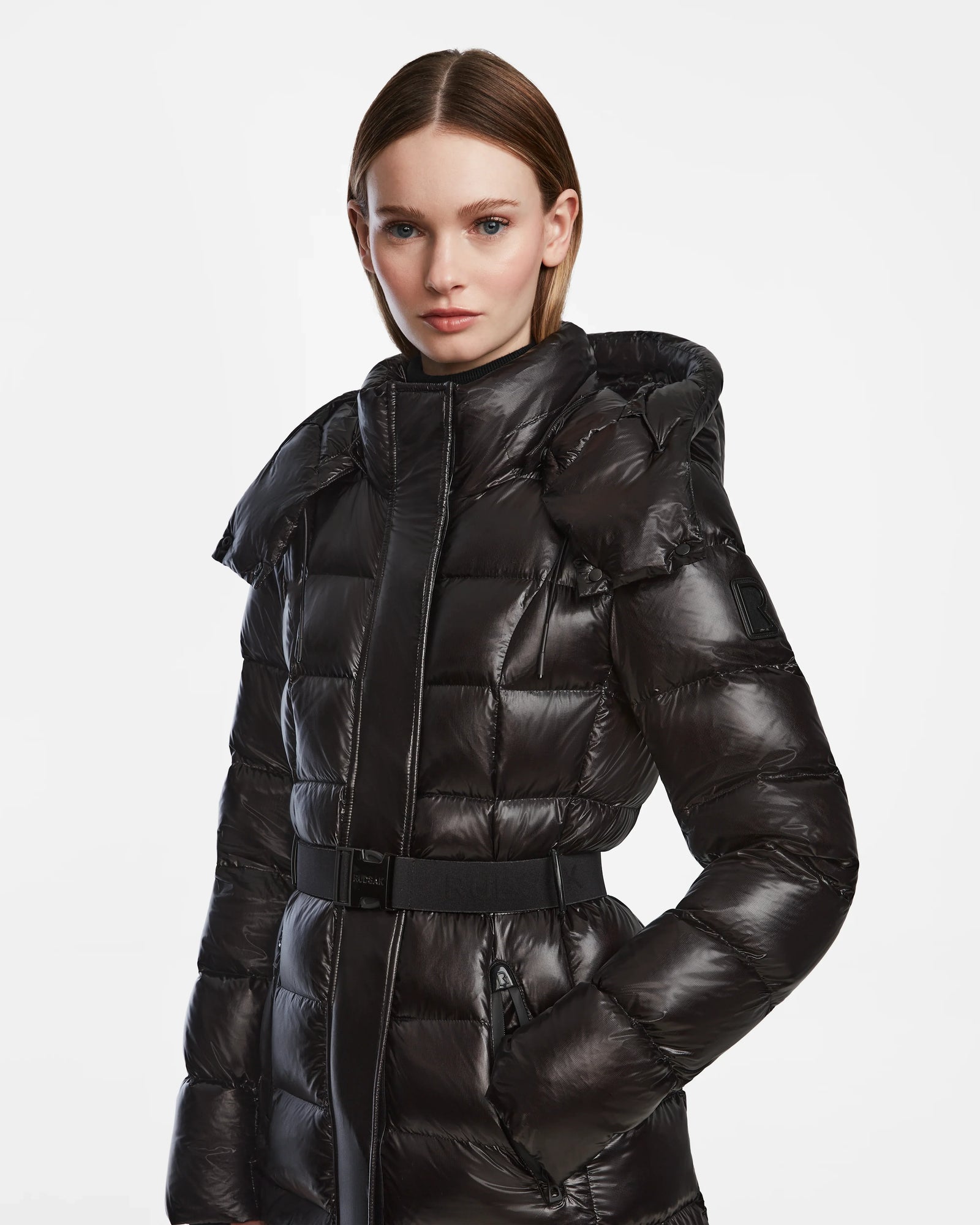 A woman with straight brown hair models the Rudsak FRIDA L-S - L124525 Black belted puffer coat with a hood, standing against a plain white background—an ideal addition to any winter wardrobe.