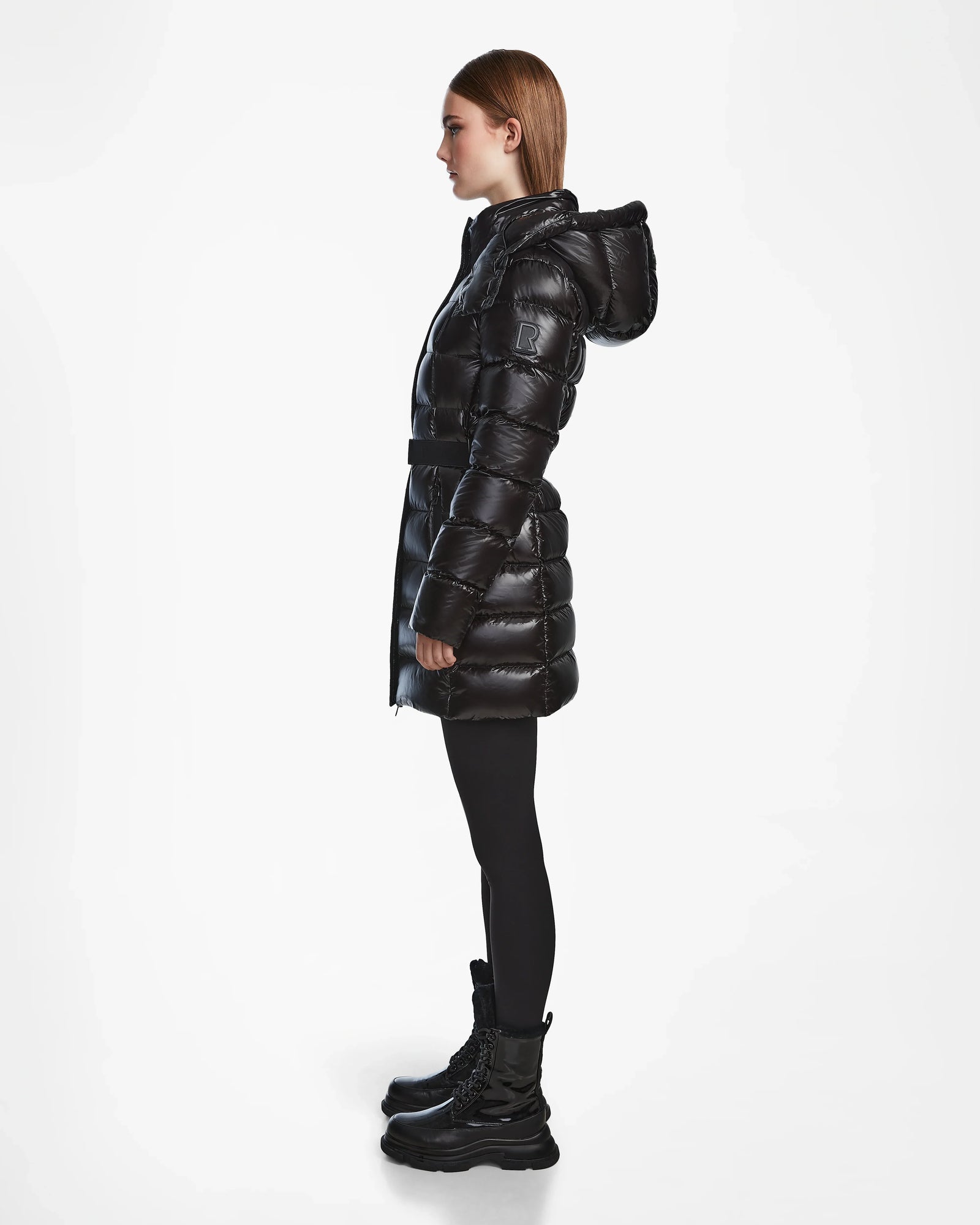 A person stands in profile wearing the Rudsak FRIDA S - L124526 Black shiny quilted down jacket with a removable hood, paired with black leggings and boots, against a plain white background.