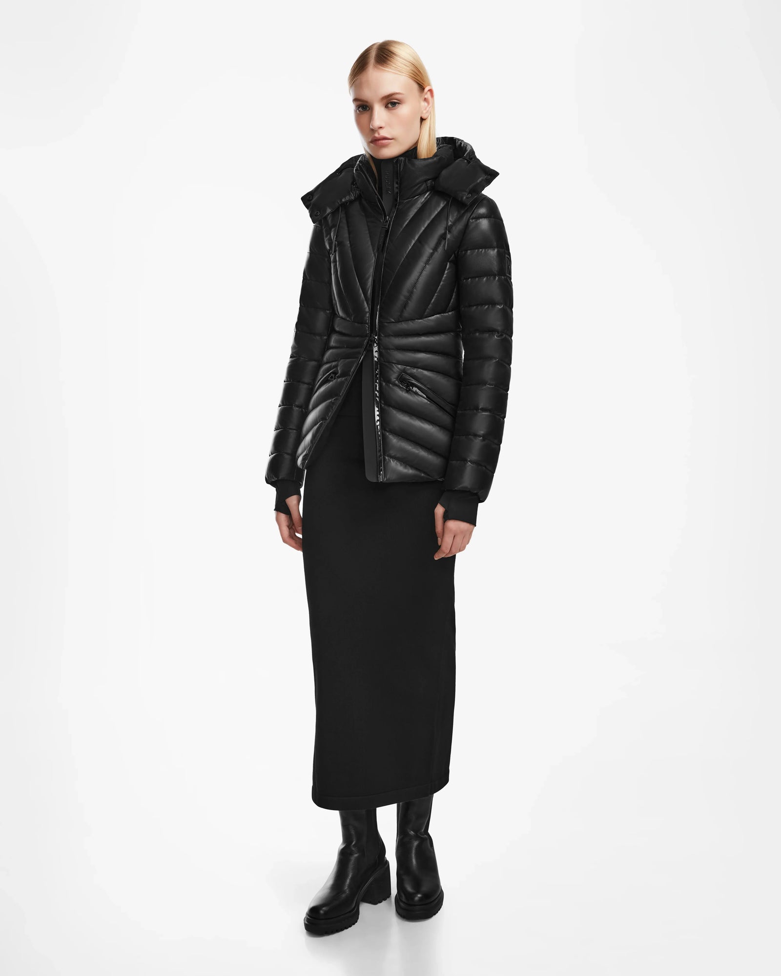 A woman with straight blonde hair and a neutral expression stands against a white background wearing the Rudsak HANNA X - 8123558 Black luxury down jacket, a long black dress, and black boots.