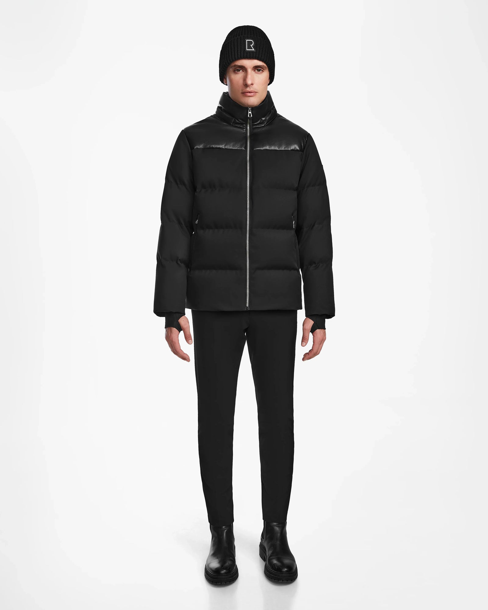 A person stands facing forward in the Rudsak JACK - M124507 Black men’s down puffer, paired with black pants, boots, and a beanie featuring a small logo, against a plain white background.