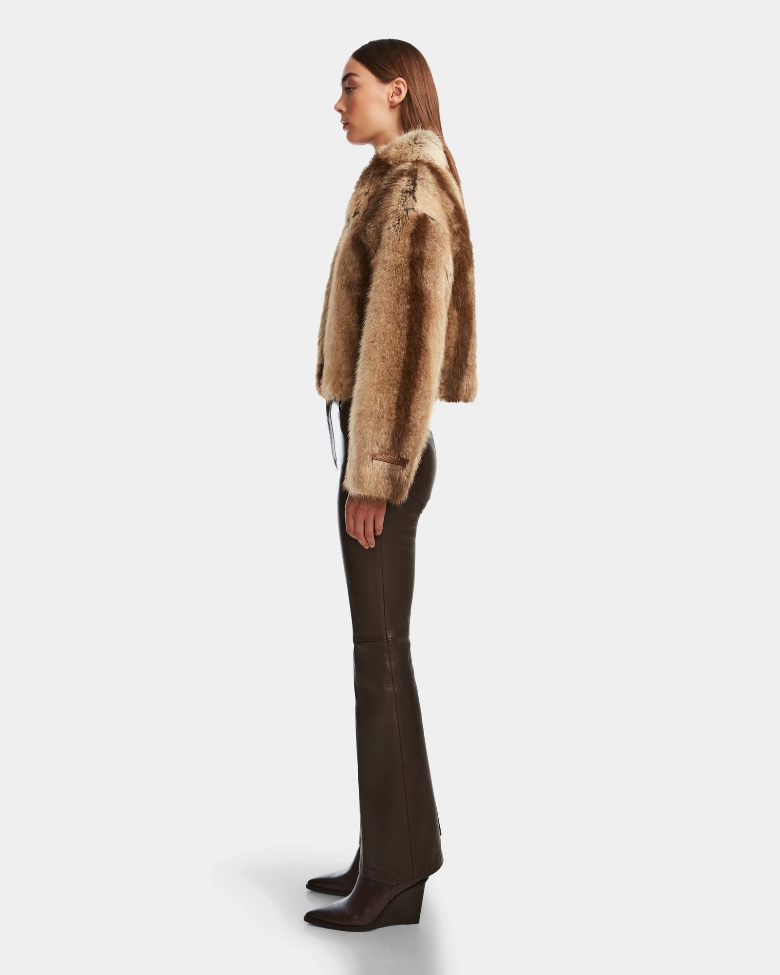 A woman in profile stands against a white background wearing the Rudsak JOSILYN - 8124792 Mocha faux fur jacket, dark brown leather pants, brown heeled boots, and her straight hair parted in the middle.