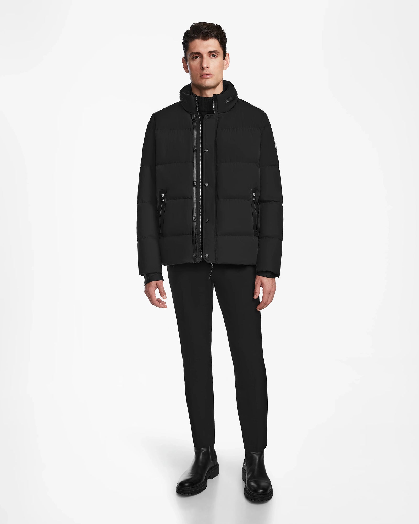 A man faces forward against a white background, wearing the Rudsak KARLS - M124505 Black mens puffer jacket with RDS-certified down, paired with black pants and boots.