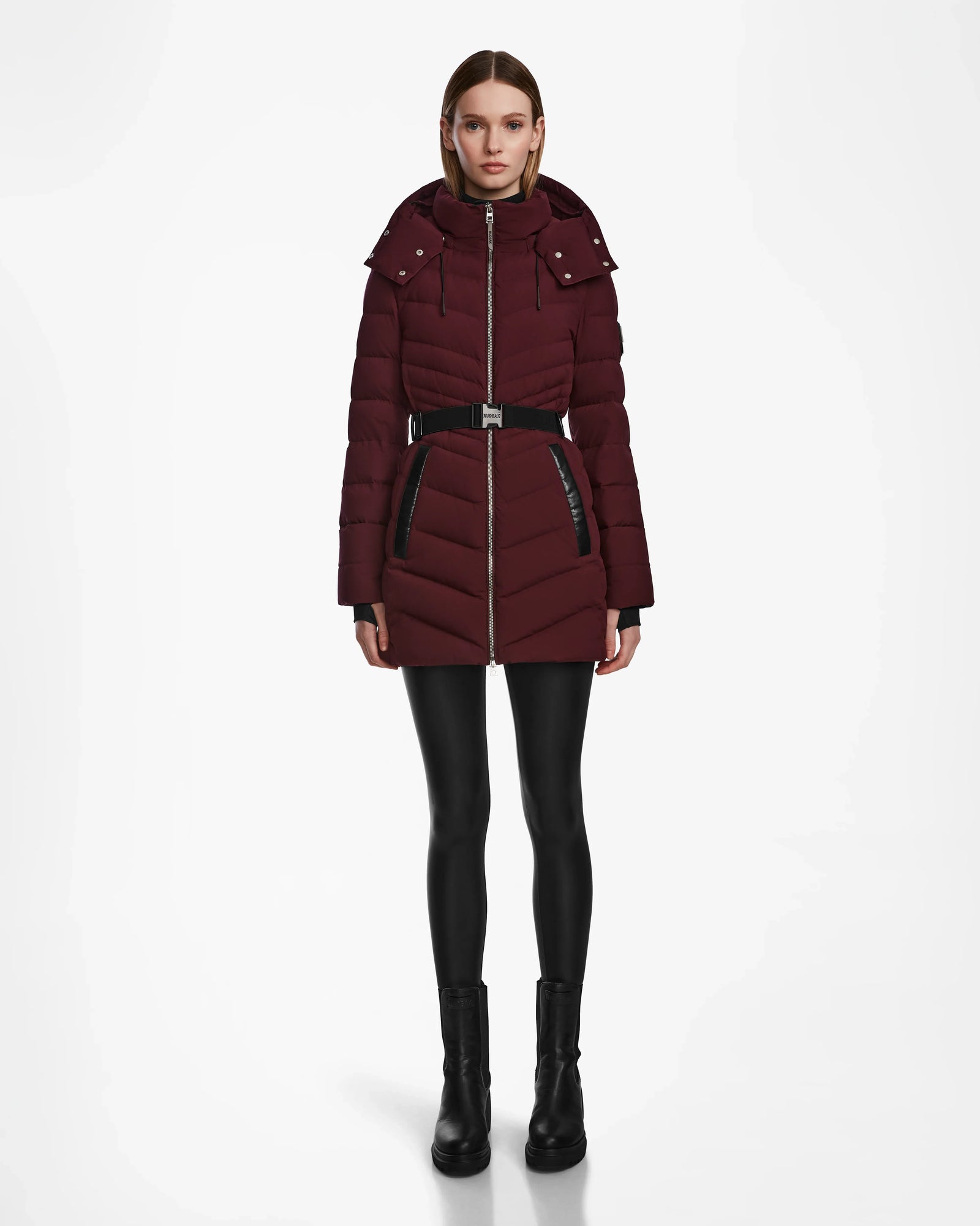 A woman stands facing forward, wearing the Rudsak MALAYA M - L123554 Wine women’s down puffer jacket, black leggings, and black ankle boots against a plain white background.