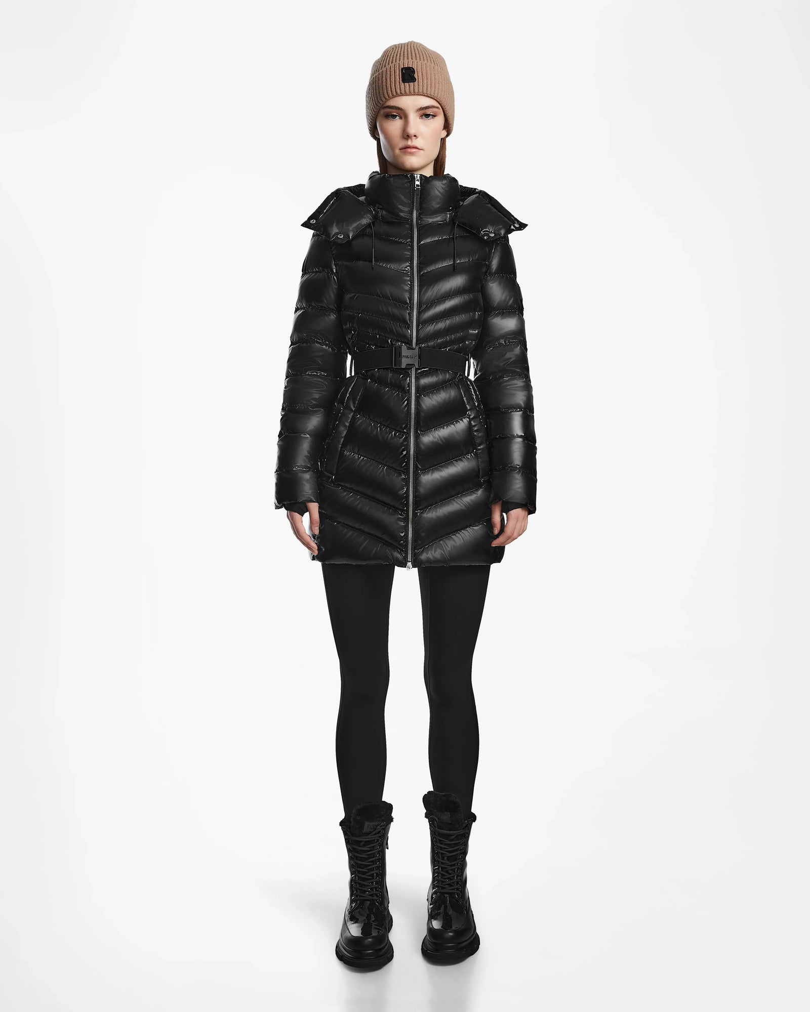 A person faces forward against a white background wearing the Rudsak MALAYA S - L123510 Black water-resistant women’s down puffer jacket, styled with black leggings, black lace-up boots, and a beige knit beanie.