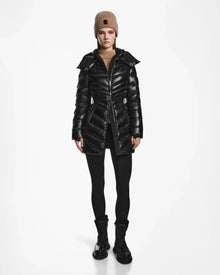 A woman wearing Rudsaks MALAYA S - L123510 Black water resistant women’s down puffer jacket with herringbone quilting, black leggings, lace-up boots, and a beige knit beanie stands facing forward against a white background.