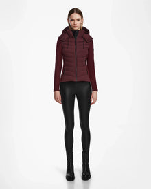 A woman stands facing forward, wearing the Rudsak MALYA M - L123547 Wine down quilted jacket, black shiny leggings, and black boots against a plain white background.