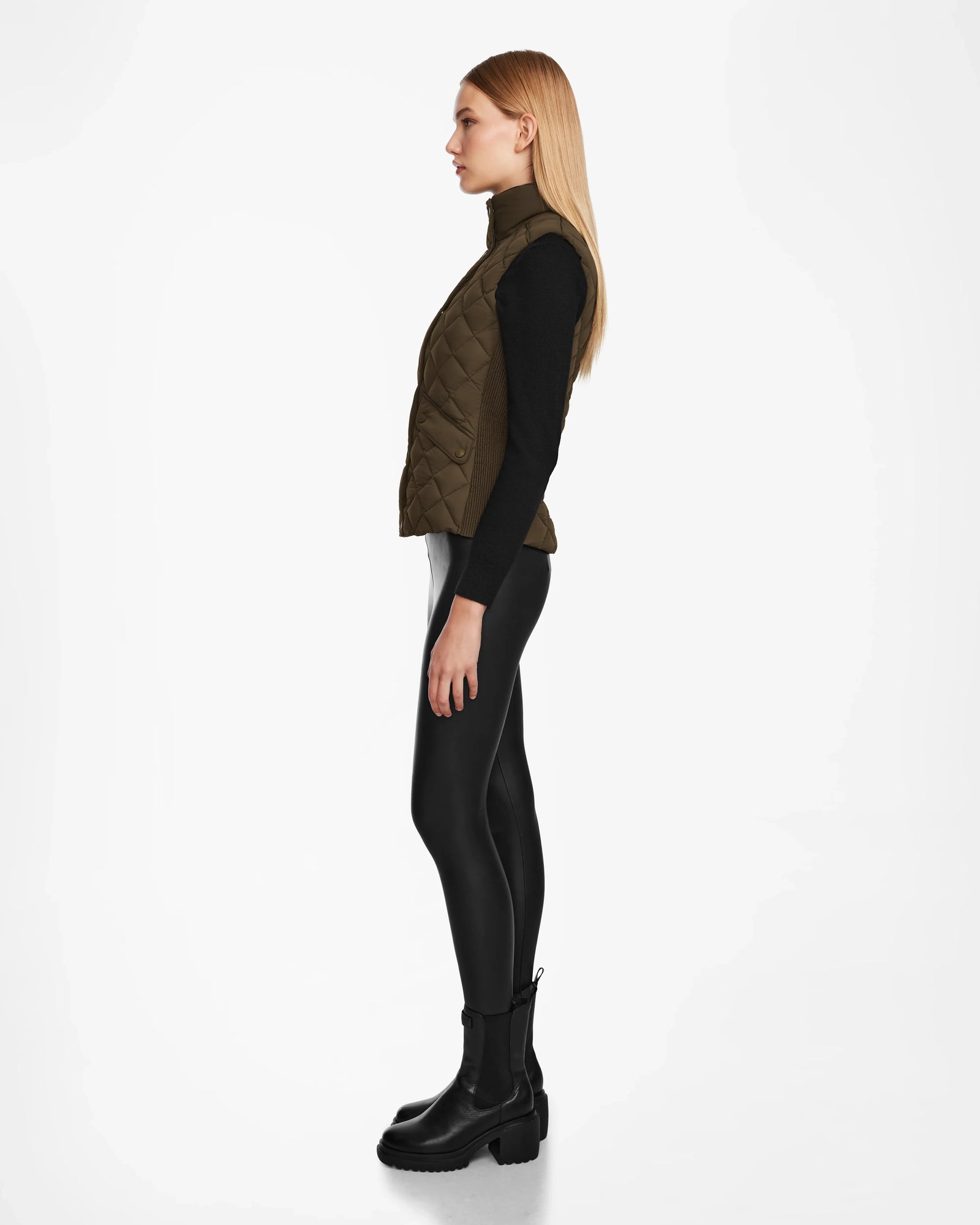 A woman with long blonde hair stands in profile against a white background, wearing the Rudsak MATILDA - L124504 Army vest over a black long-sleeve top, shiny black leggings, and black boots.