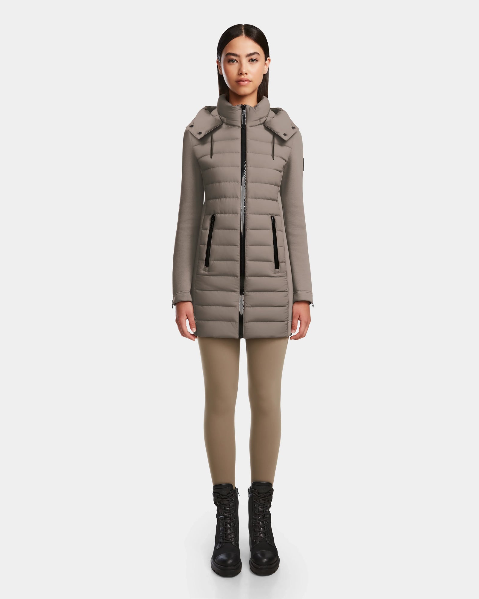 A woman faces forward against a white background, wearing the Rudsak MERINA M - 8124030 Cement taupe quilted down jacket with a high collar, tan leggings, and black lace-up boots.