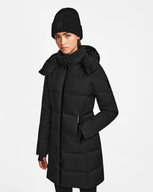 A woman wears the Rudsak MIRIAM - L122523 BLACK womens down puffer coat with hood, plus a black beanie and gloves, standing against a white background. She has her left hand in her pocket. The coat is filled with RDS-certified down.