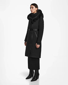 A woman stands in a studio wearing the Rudsak MUSA - L124534 Black women’s long down puffer with a fur-trimmed hood, belted waist, and zippered pockets. She pairs it with a black skirt and pointed ankle boots, facing slightly to the side.