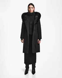 A woman faces forward wearing the Rudsak MUSA - L124534 Black long down puffer with a fur-trimmed hood, paired with a black skirt, black boots, and a high-neck layer, set against a plain white background.