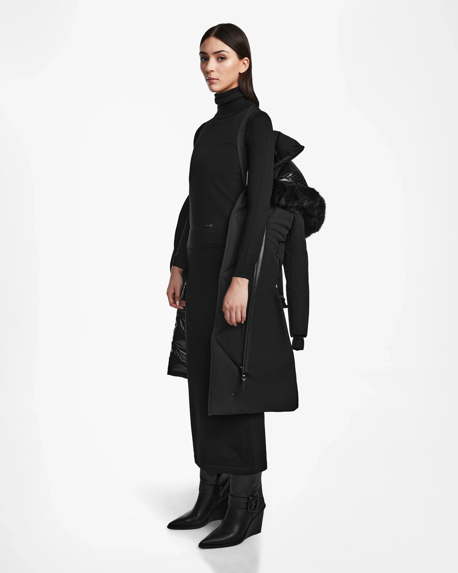 A woman stands in profile against a white background, dressed in black and holding the Rudsak MUSA - L124534 Black sustainable parka with a furry hood draped over her shoulder.