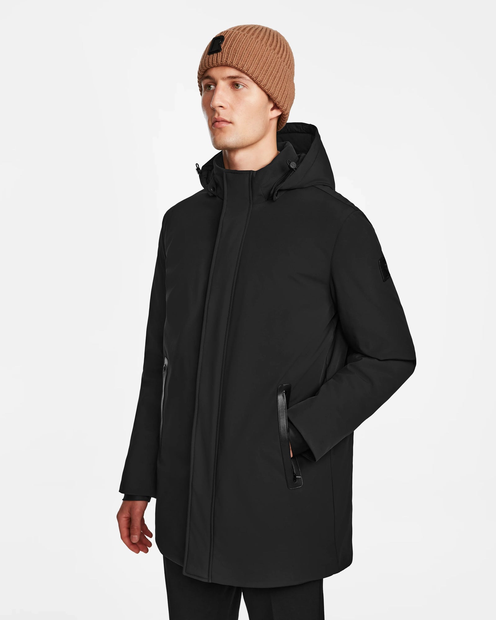 A man wears the Rudsak NATAN - M124519 Black parka and a brown knit beanie, standing against a plain white background and looking slightly to the side with a neutral expression.