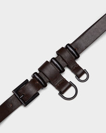 The PREYA - 8324508 Espresso belt by Rudsak is a dark brown leather accessory with a black rectangular buckle, two black D-rings, and three leather loops, offering a modern utilitarian look displayed against a light grey background.