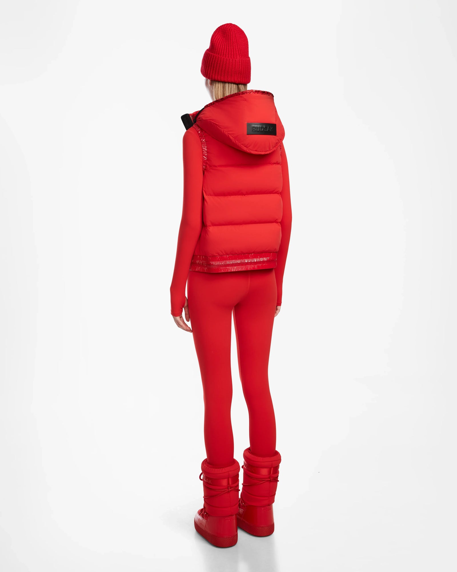 A person in the Rudsak RAQUEL M - 8124538 Flame Red reversible ski vest, beanie, long-sleeve top, leggings, and boots stands facing away against a plain white background.