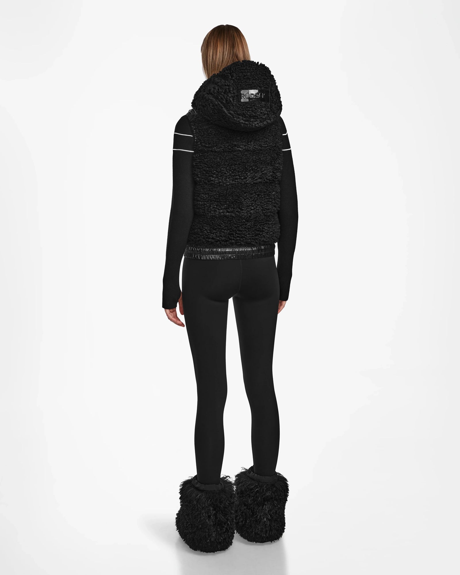 A person faces away, wearing the RAQUEL S - 8124526 Black ski vest by Rudsak, paired with a black long-sleeve top, black leggings, and large black furry boots against a plain white background.