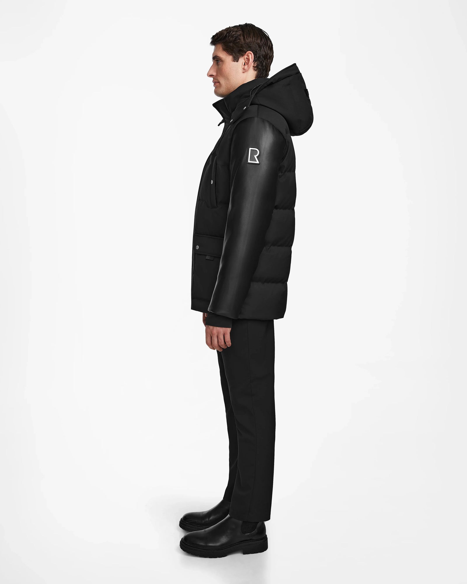 A man stands in profile wearing the Rudsak RICKY R - M123506 Black men's quilted down parka with recycled leather sleeves, paired with black pants and boots, against a plain white background.