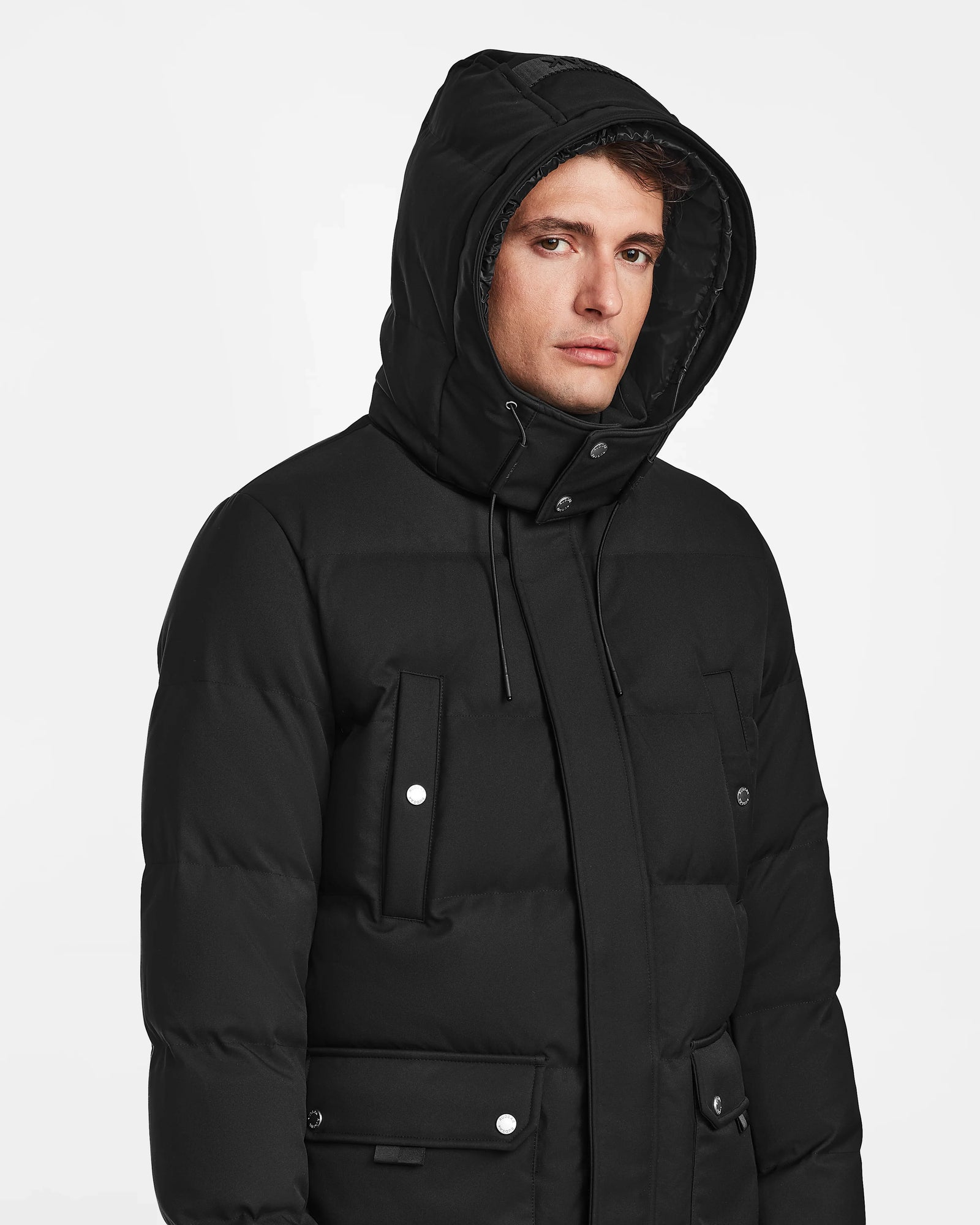 A person wearing the Rudsak RICKY - M123502 Black men's quilted down parka with the hood up, looking slightly to the side against a plain white background.