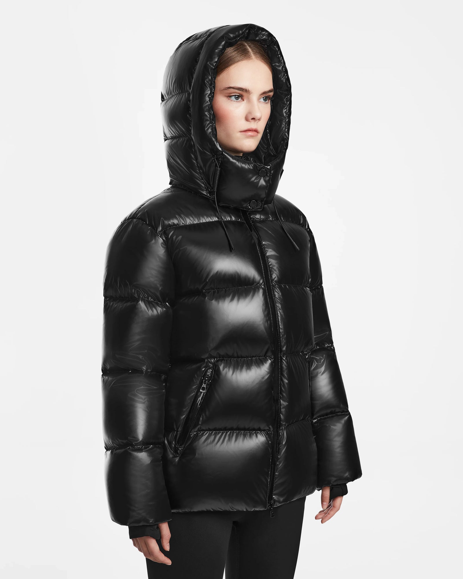 A woman wears the Rudsak RITA S - L124520 Black hooded down puffer jacket with horizontal quilting, zipped up, standing against a plain white background.