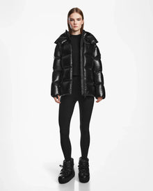 A person wearing the Rudsak RITA S - L124520 Black weather-resistant puffer jacket stands against a plain white background in black leggings and boots, with straight brown hair and a neutral expression, arms relaxed at their sides.