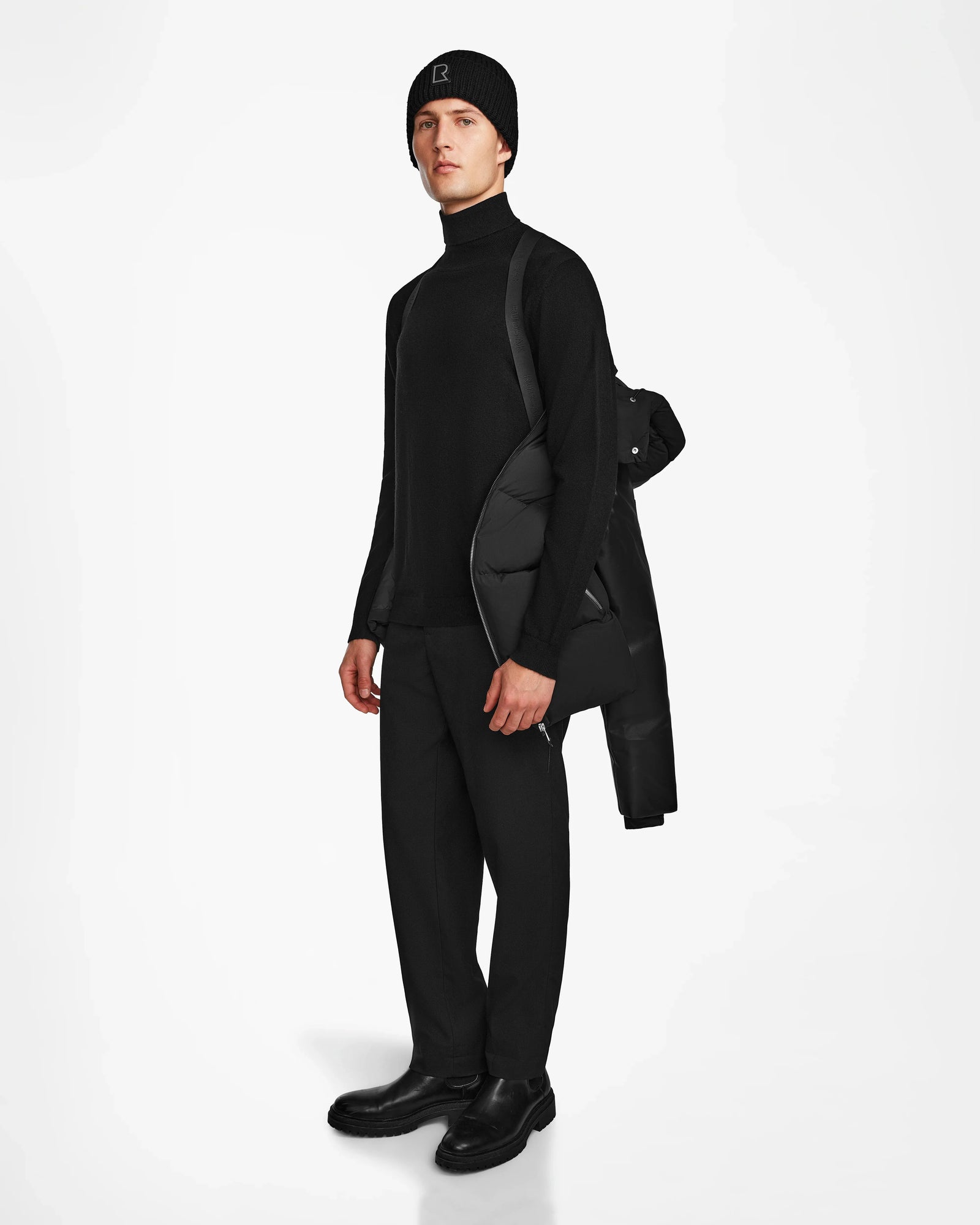 A person models Rudsaks RONNY M - M124513 Black winter puffer, crafted from recycled materials, draped over one shoulder while dressed in all black, standing against a plain white background.