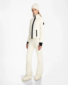 A woman stands against a white background wearing the Rudsak ROSALIE - 8124784 Iceland women's fitted ski jacket with black zipper details, matching white pants, a white beanie, black gloves, and platform boots—perfect for stylish winter adventures.