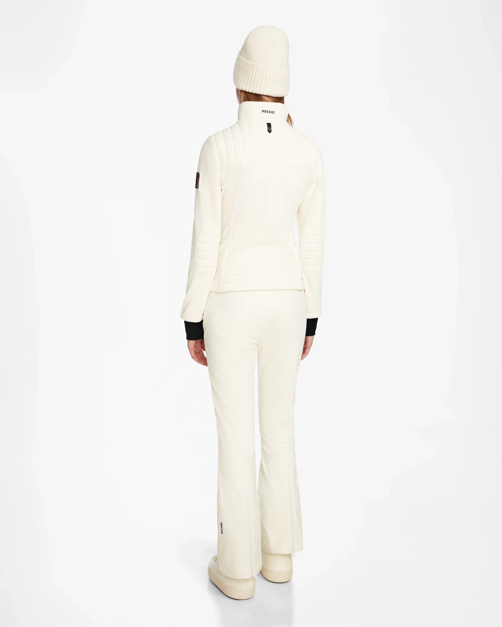 A person faces away in an all-white Rudsak ROSALIE - 8124784 Iceland winter look, featuring a women’s fitted ski jacket with RECCO rescue tech, flared pants, and beanie, set against a plain white background.