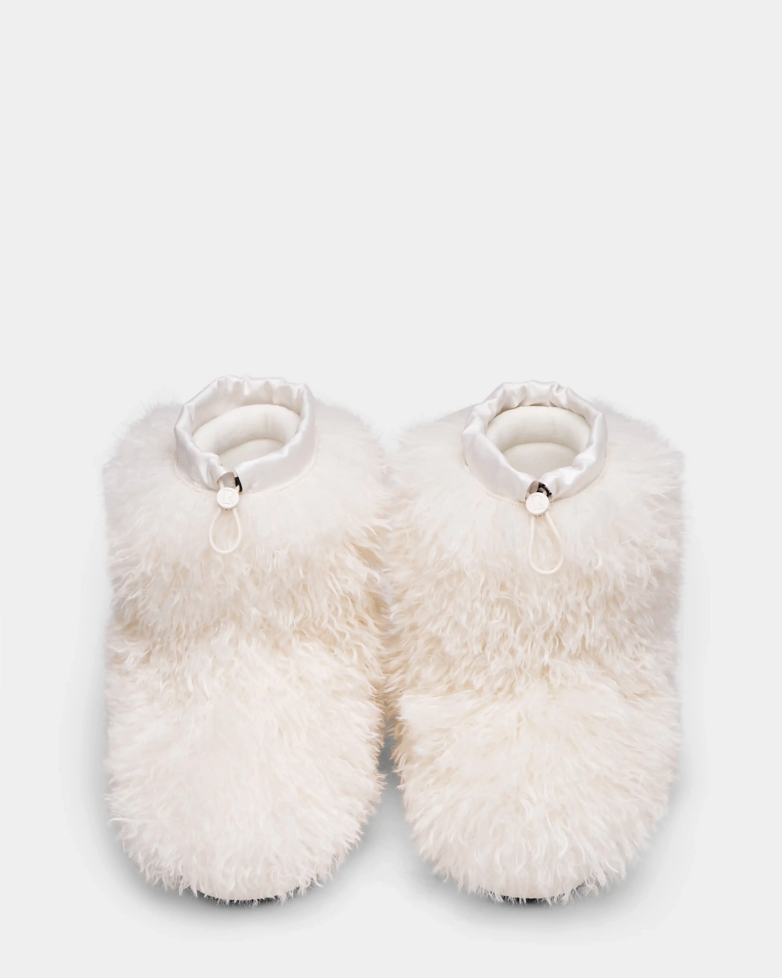 The ROSSY - 8224518 Iceland slippers by Rudsak are fluffy white with a soft, shaggy Mongolian fur-like texture, satin-like ankle trim, and feature a small decorative loop and bead on each slipper against a plain light background.
