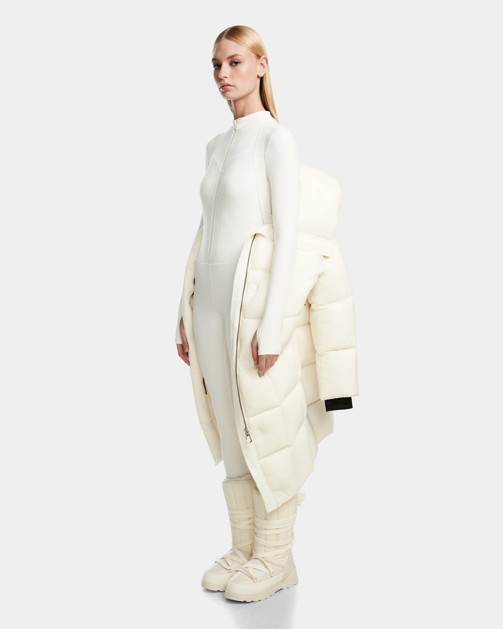 A woman wears the Rudsak SASH - 8124536 Iceland oversized women’s down puffer coat draped over her arms, styled with a fitted white bodysuit and matching puffy boots, set against a plain light gray background.